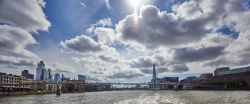 This urban landscape photograph captures the Millennium Bridge spanning the River Thames in London during a summer morning. The viewpoint is from the river itself, looking towards the bridge, with the sun visible high behind cloud cover and reflecting on the water. Notable examples of contemporary London architecture are present, including The Shard centrally located in the background, the distinct city skyline to the left, and riverside buildings along the Thames embankment. The Tate Modern is seen on the right, contributing to the mix of modern and historic architecture surrounding the river. The photograph highlights London's blend of urban development and iconic landmarks along the Thames under a partly cloudy summer sky.